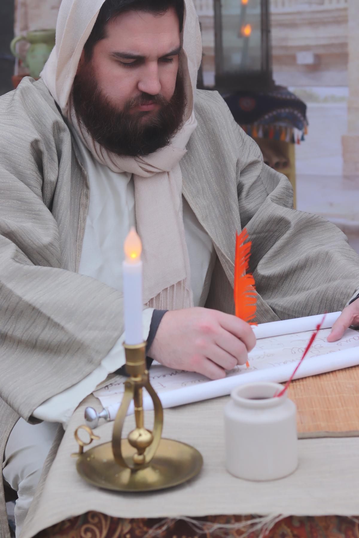 A scribe in robes writes by candlelight on a scroll with a quill.