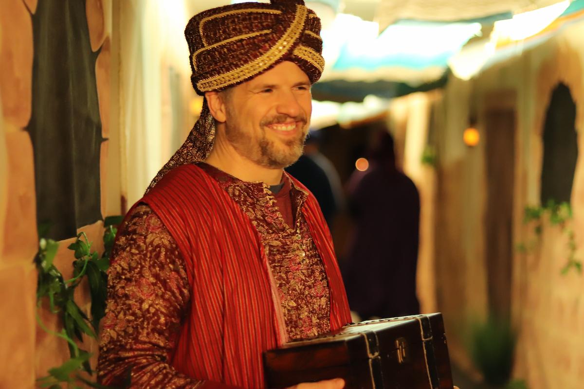 A wise man in colorful robes smiles warmly while holding a treasure chest.